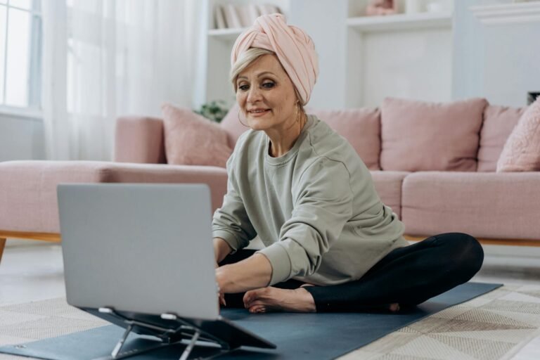 Senior woman using a laptop for an online yoga class, sitting indoors on a yoga mat with a relaxed expression.