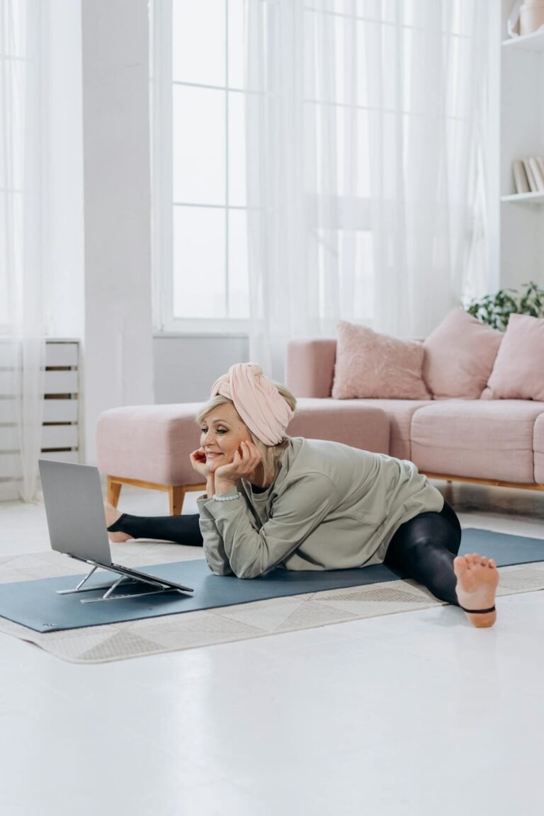 Senior woman using a laptop while practicing yoga at home in a bright living room.